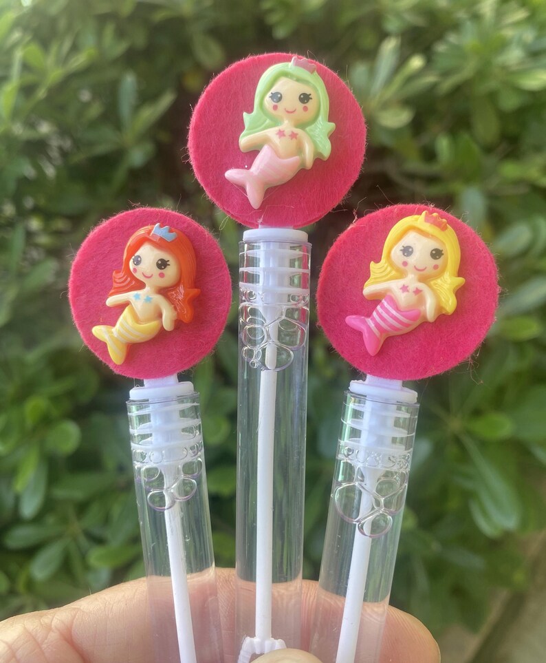 Op de afbeelding: Drie bellenblaasstokken met zeemeermin-toppers. Elke toverstaf heeft een doorzichtige plastic buis met een witte toverstaf en een roze vilten cirkel met een zeemeerminfiguur. De zeemeerminnen hebben verschillende haar- en staartkleuren: groen, oranje en geel.