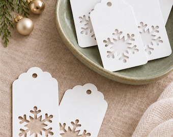 May include: A collection of white rectangular gift tags with scalloped tops and a circular hole for hanging. Each tag features a die-cut snowflake design in the center. Several tags are arranged inside a shallow green bowl.