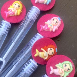 Op de afbeelding: Bellenblazers met roze vilten cirkels en zeemeerminversieringen. De zeemeerminnen hebben verschillende haarkleuren: geel, blauw, roze, rood en groen. De bellenblazers zijn van helder plastic met een witte toverstaf erin.