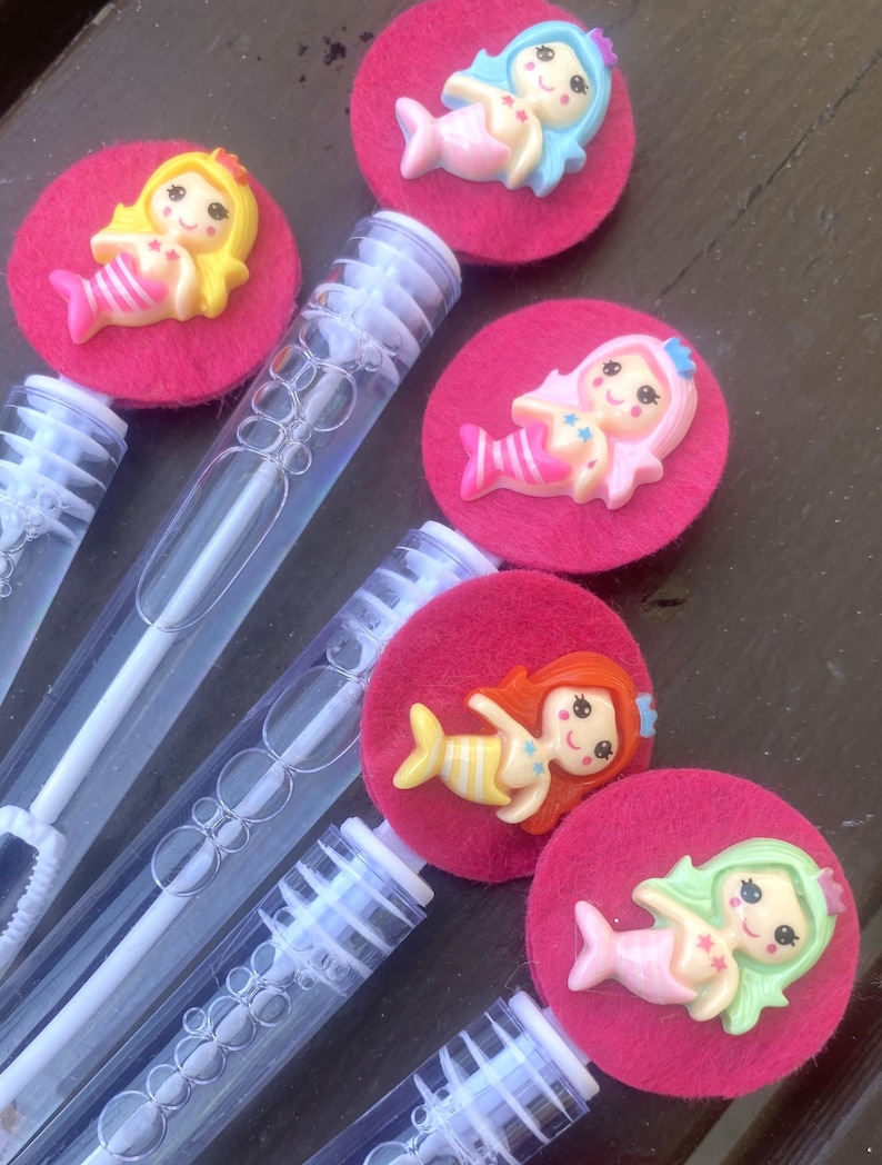 Op de afbeelding: Bellenblazers met roze vilten cirkels en zeemeerminversieringen. De zeemeerminnen hebben verschillende haarkleuren: geel, blauw, roze, rood en groen. De bellenblazers zijn van helder plastic met een witte toverstaf erin.