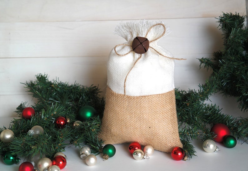 May include: A white and burlap Christmas gift bag with a brown bell tied with twine. The bag is surrounded by green pine branches and red, green, and white ornaments.