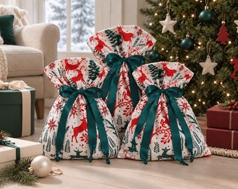 May include: Three fabric gift bags in varying sizes, featuring a white background with a repeating pattern of red reindeer and green pine trees. Each bag is cinched at the top with a dark green satin ribbon tied into a decorative bow.