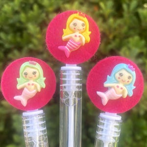 Op de afbeelding: Drie bellenblaasstokken met zeemeermin-toppers. Elke toverstaf heeft een heldere plastic fles met een roze vilten cirkel en een zeemeerminfiguur. De zeemeerminnen hebben verschillend gekleurd haar en staarten.