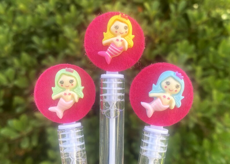 Op de afbeelding: Drie bellenblaasstokken met zeemeermin-toppers. Elke toverstaf heeft een heldere plastic fles met een roze vilten cirkel en een zeemeerminfiguur. De zeemeerminnen hebben verschillend gekleurd haar en staarten.