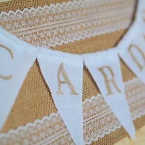 May include: White fabric bunting with the word "CARDS" written in gold lettering. The bunting is strung on a burlap background with white lace.