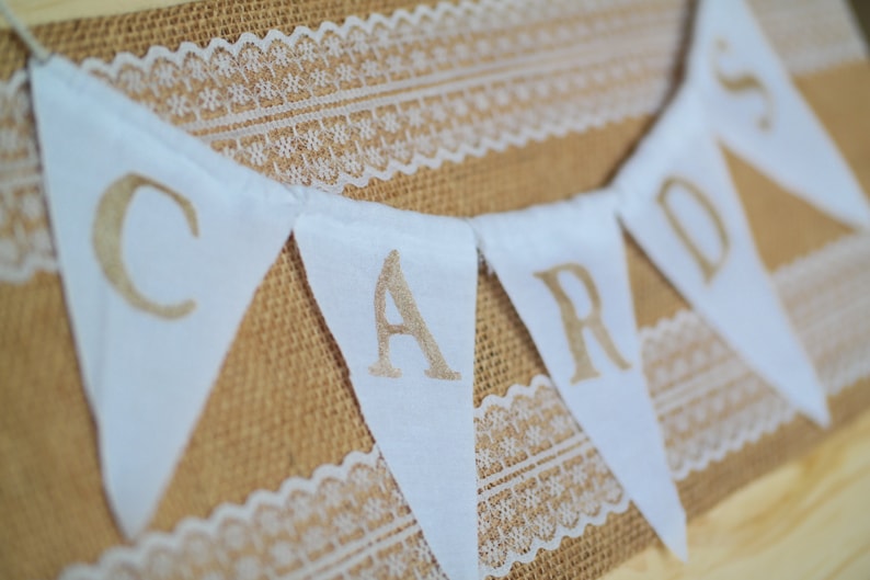 May include: White fabric bunting with the word "CARDS" written in gold lettering. The bunting is strung on a burlap background with white lace.