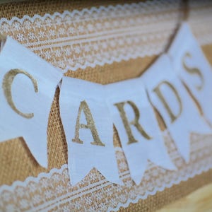 May include: White fabric banner with gold lettering that reads "Cards" hanging on a burlap background with white lace trim.