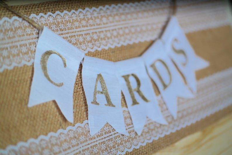 May include: White fabric banner with gold lettering that reads "Cards" hanging on a burlap background with white lace trim.