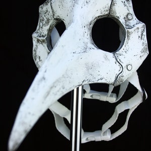 Puede incluir: Una m&aacute;scara blanca de m&eacute;dico de la peste con un pico largo y curvo y agujeros para los ojos. La m&aacute;scara tiene una apariencia envejecida y desgastada con acentos oscuros y se asegura con correas y hebillas blancas. La m&aacute;scara se exhibe sobre un soporte plateado sobre un fondo negro.