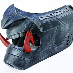 Könnte beinhalten: Eine detailreiche Maske in Silber und Rot mit dem Wort "ARASAKA" auf der Seite. Die Maske hat eine strukturierte, metallische Oberfläche und ein scharfes, kantiges Design.