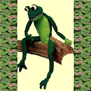 May include: A green crocheted frog sitting on a brown branch. The frog has large, round eyes and a wide smile. The frog is surrounded by a border of pink water lilies and green lily pads.