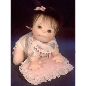 May include: A handmade cloth doll with brown hair, wearing a pink and white dress and a white bib with a colorful pattern. The doll is lying on a pink and white pillow with lace trim.
