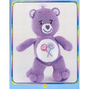 May include: A purple crocheted teddy bear with a white heart on its chest and two pink and blue lollipops. The bear has a white face with black eyes and a pink nose.