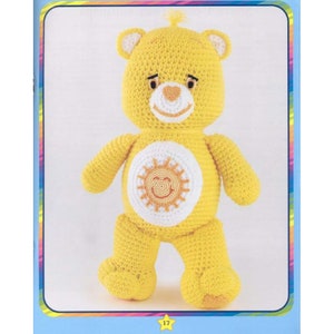 May include: A yellow crocheted Care Bear with a white sun with a smiling face on its chest. The bear has a pink heart nose and black eyes.