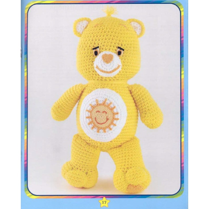 May include: A yellow crocheted Care Bear with a white sun with a smiling face on its chest. The bear has a pink heart nose and black eyes.
