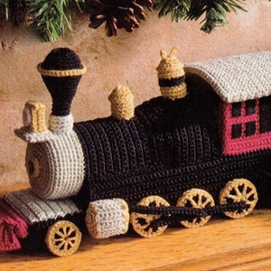 Peut inclure: Un train jouet en crochet avec des détails noirs, blancs, gris et rouges. Le train a un corps noir, des roues blanches et un wagon de queue rouge. Le train est décoré d'une cheminée jaune et d'une cloche jaune.