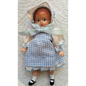May include: A small doll with reddish-brown hair wearing a light blue and white gingham dress with puffy sleeves and a matching bonnet tied with a light blue ribbon. The doll has painted facial features and wears white socks with black shoes.