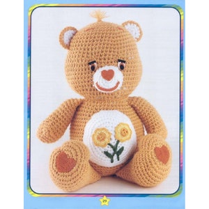 May include: A crocheted Care Bear plush toy with a yellow flower design on its belly. The bear is sitting with its arms crossed and has a heart-shaped nose.