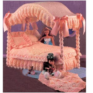 May include: A peach-colored, crocheted doll bed with a canopy, pillows, and a bedspread. The bed has a ruffled skirt and is decorated with pink bows. Two dolls and small teddy bears are on the bed and floor.