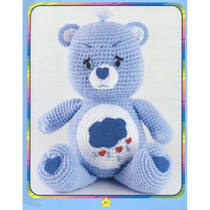 May include: A crocheted blue Care Bear with a white belly and a blue cloud with red raindrops on it. The bear has a heart-shaped nose and a grumpy expression.