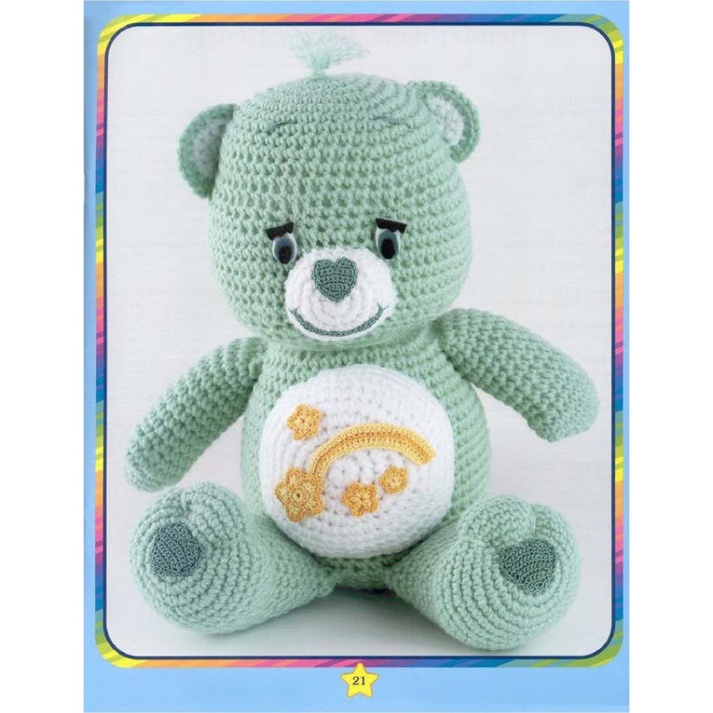 May include: A crocheted green teddy bear with a white belly featuring a yellow rainbow and stars. The bear has a heart-shaped nose and black eyes.
