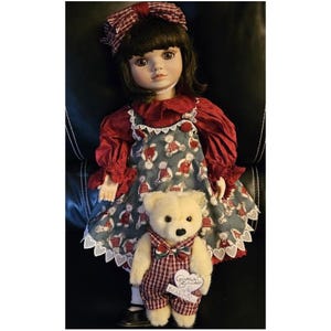 May include: A porcelain doll with dark hair and a red bow, wearing a red dress with a blue and white teddy bear print apron. A small, cream-colored teddy bear in red plaid overalls is held in front of the doll.