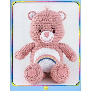 May include: A pink crocheted Care Bear with a rainbow on its belly. The bear has a white face with a pink nose and black eyes.