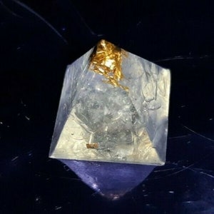 Può includere: Un cristallo trasparente a forma di piramide con una parte superiore dorata. Il cristallo è posto su uno sfondo scuro, con un riflesso viola alla base. Le sfaccettature del cristallo catturano la luce, creando un effetto scintillante.