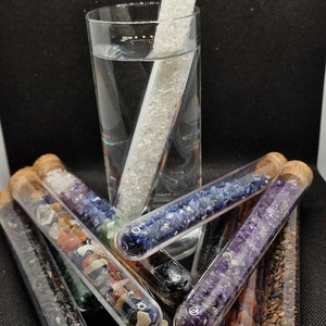 Könnte beinhalten: Eine Sammlung von Glasröhrchen, gefüllt mit bunten Edelsteinen und Kristallen, jeweils mit einem Korken verschlossen. Ein Röhrchen befindet sich in einem Glas Wasser. Die Edelsteine umfassen Amethyst, blauen Sodalith und Bergkristall.