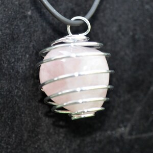 Può includere: Una collana con pendente in quarzo rosa. La pietra sferica rosa chiaro è racchiusa in una gabbia di filo argentato e pende da un cordino grigio scuro. Il pendente è un gioiello in pietra naturale.
