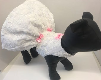 May include: White dog dress with a ruffled skirt and pink bows. The dress is made of a soft, white fabric with a rose pattern.
