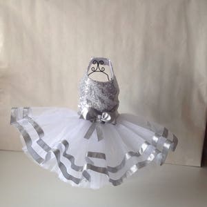 May include: A white tulle tutu skirt with silver ribbon trim and a silver sequined bodice. The skirt is on a white mannequin.