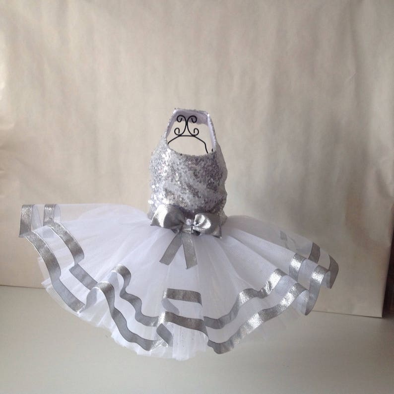 May include: A white tulle tutu skirt with silver ribbon trim and a silver sequined bodice. The skirt is on a white mannequin.
