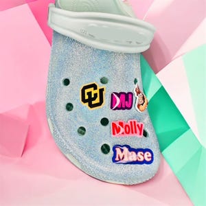 May include: A light blue glittery clog shoe with multiple decorative charms. The charms include the letters "CU", "DMJ", and the name "Molly" and "Mase". The shoe has a white strap and is set against a pink and green background.