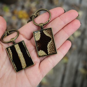 May include: Two keyrings with rectangular metal frames. Each frame contains a different pattern of brown and white snake skin.