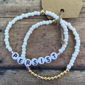 May include: Two white beaded bracelets with gold beads. The top bracelet has the word "PERSIST" spelled out in black letters. The bottom bracelet has a tag attached that says "hold on tight".