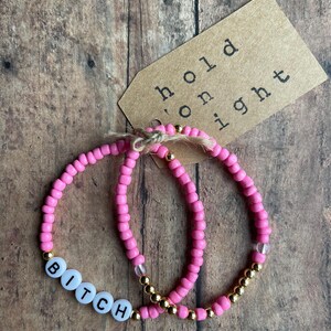 May include: Two pink beaded bracelets with gold beads. One bracelet has the word "BITCH" spelled out in white letters.