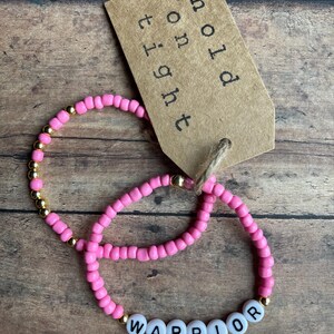 May include: Two pink beaded bracelets with gold beads. One bracelet has the word "WARRIOR" spelled out in white beads. A brown tag with the text "hold on tight" is in the background.