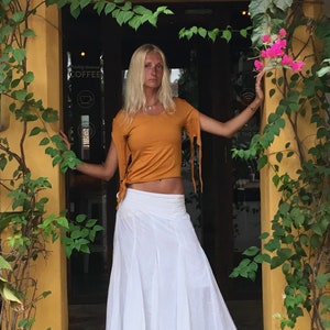 May include: A woman wearing a mustard yellow crop top with long, dangling ties and a white maxi skirt. The outfit is paired with black sandals. The background includes green foliage, pink flowers, and a yellow doorway.