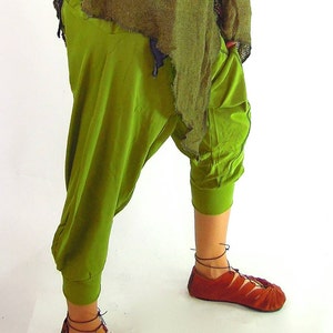 May include: A person wearing a green outfit with a distressed, olive green top and loose-fitting, olive green pants. The outfit includes red, lace-up sandals.