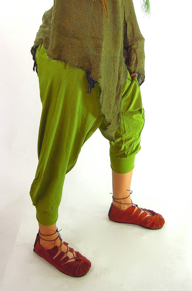May include: A person wearing a green outfit with a distressed, olive green top and loose-fitting, olive green pants. The outfit includes red, lace-up sandals.