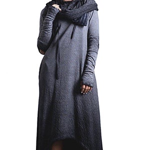 Puede incluir: Vestido largo gris con capucha, mangas largas y bajo asim&eacute;trico. El vestido presenta una tela texturizada y una bufanda a juego. La modelo lleva botas negras.