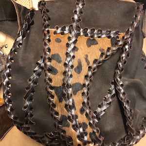 May include: A dark brown leather shoulder bag with a flap closure. The bag features a central panel of animal print fur, framed by braided leather trim. The strap is also dark brown leather.