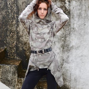 May include: A long-sleeved hooded tunic featuring a grey and beige digital camouflage pattern. The garment includes two diagonal zips at the waist and is worn with a black belt featuring silver studs and a large ornate silver-toned metal buckle.