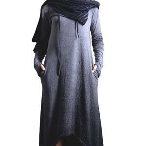 Puede incluir: Un vestido largo con capucha gris con mangas largas y bolsillos. El vestido tiene una tela texturizada y un dobladillo alto-bajo. Se usa una bufanda y una capucha negras con el vestido. La modelo lleva botines negros.