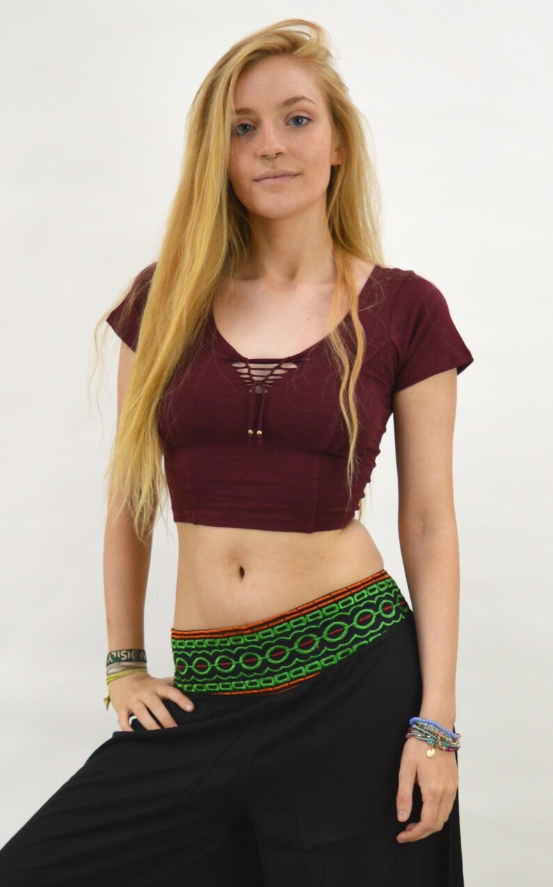 May include: A maroon crop top with a lace-up detail in the front and short sleeves. The top is paired with black pants with a green and orange embroidered waistband.