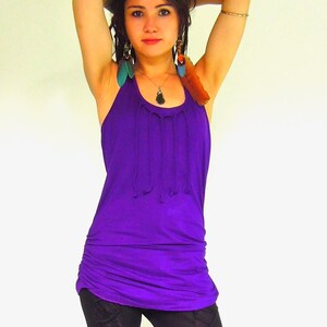 May include: A woman wearing a purple tank top with a gathered neckline and black leggings. She is wearing feather earrings and a necklace with a pendant.