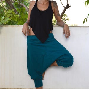 May include: A woman wearing a black tank top and teal harem pants. The tank top has a unique asymmetrical design. The pants are a vibrant teal color and have a relaxed, comfortable fit. The woman is standing barefoot.