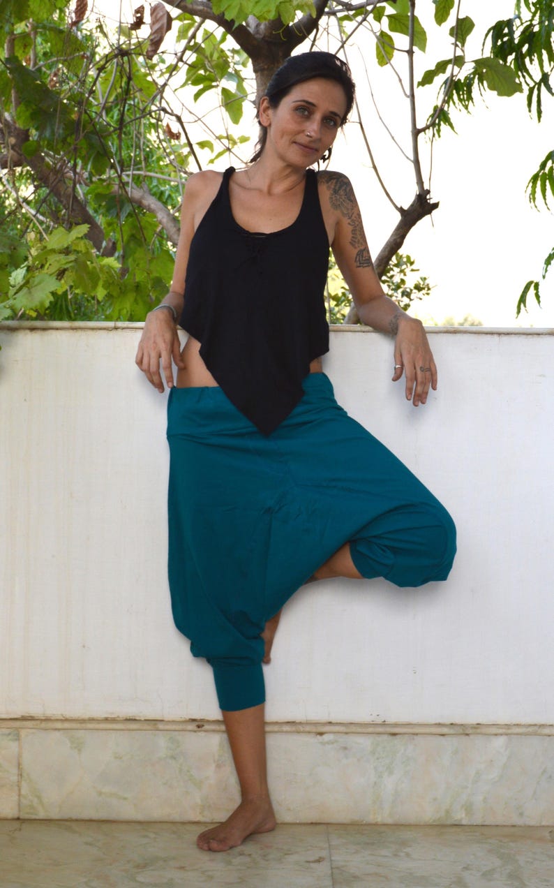 May include: A woman wearing a black tank top and teal harem pants. The tank top has a unique asymmetrical design. The pants are a vibrant teal color and have a relaxed, comfortable fit. The woman is standing barefoot.