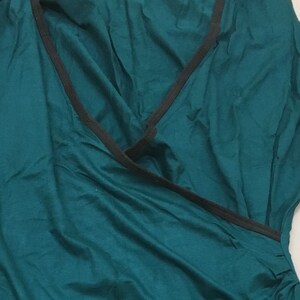 May include: A teal-colored sleeveless top with a wrap-style design. The neckline and edges are trimmed with a thin black band. The fabric has a soft, flowing appearance.